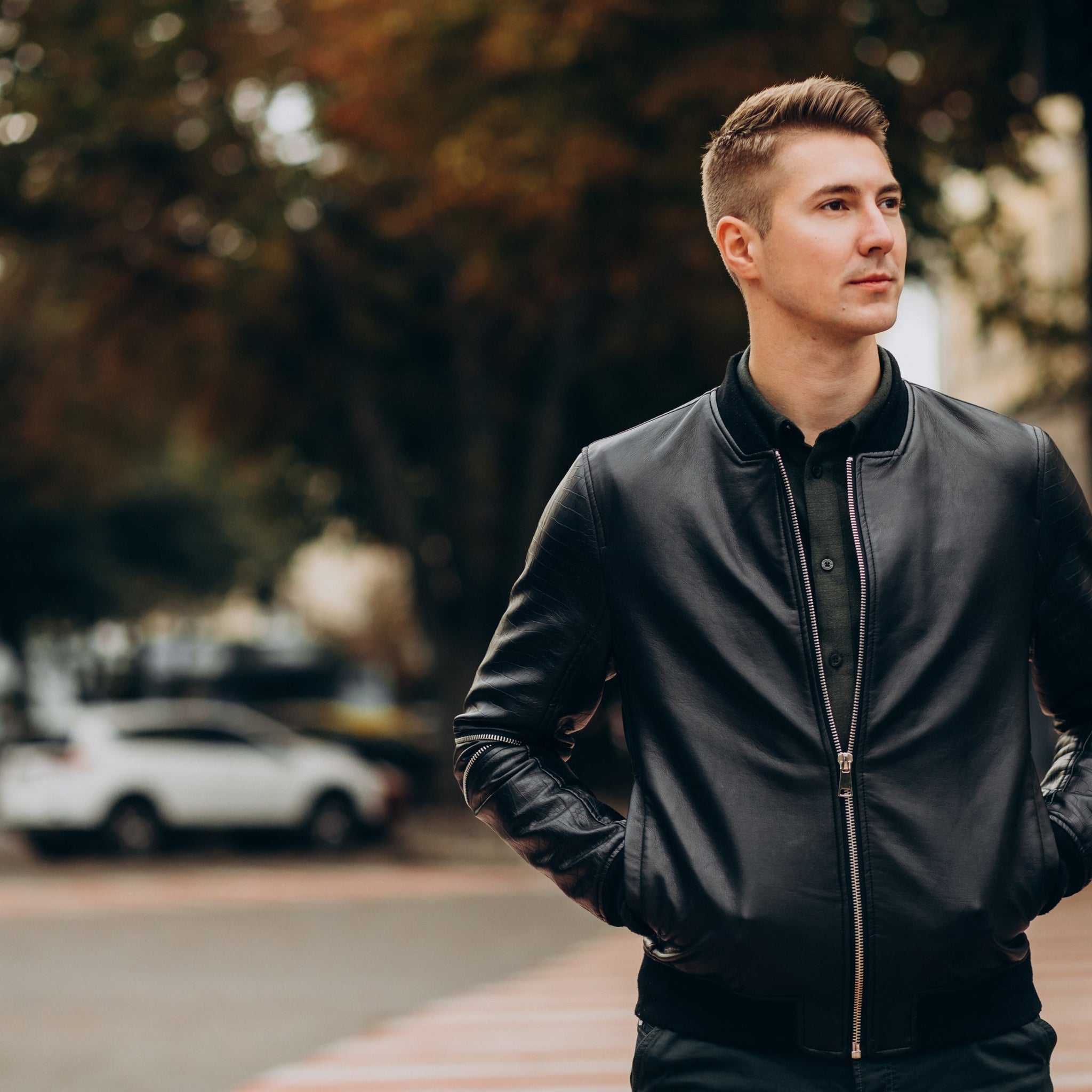 Man wearing a black leather bomber jacket, posing solo in a modern city backdrop.