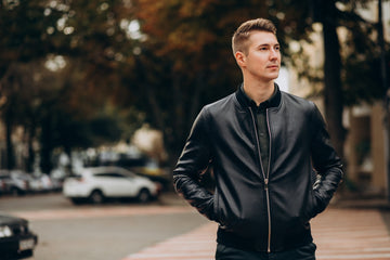Man wearing a black leather bomber jacket, posing solo in a modern city backdrop.